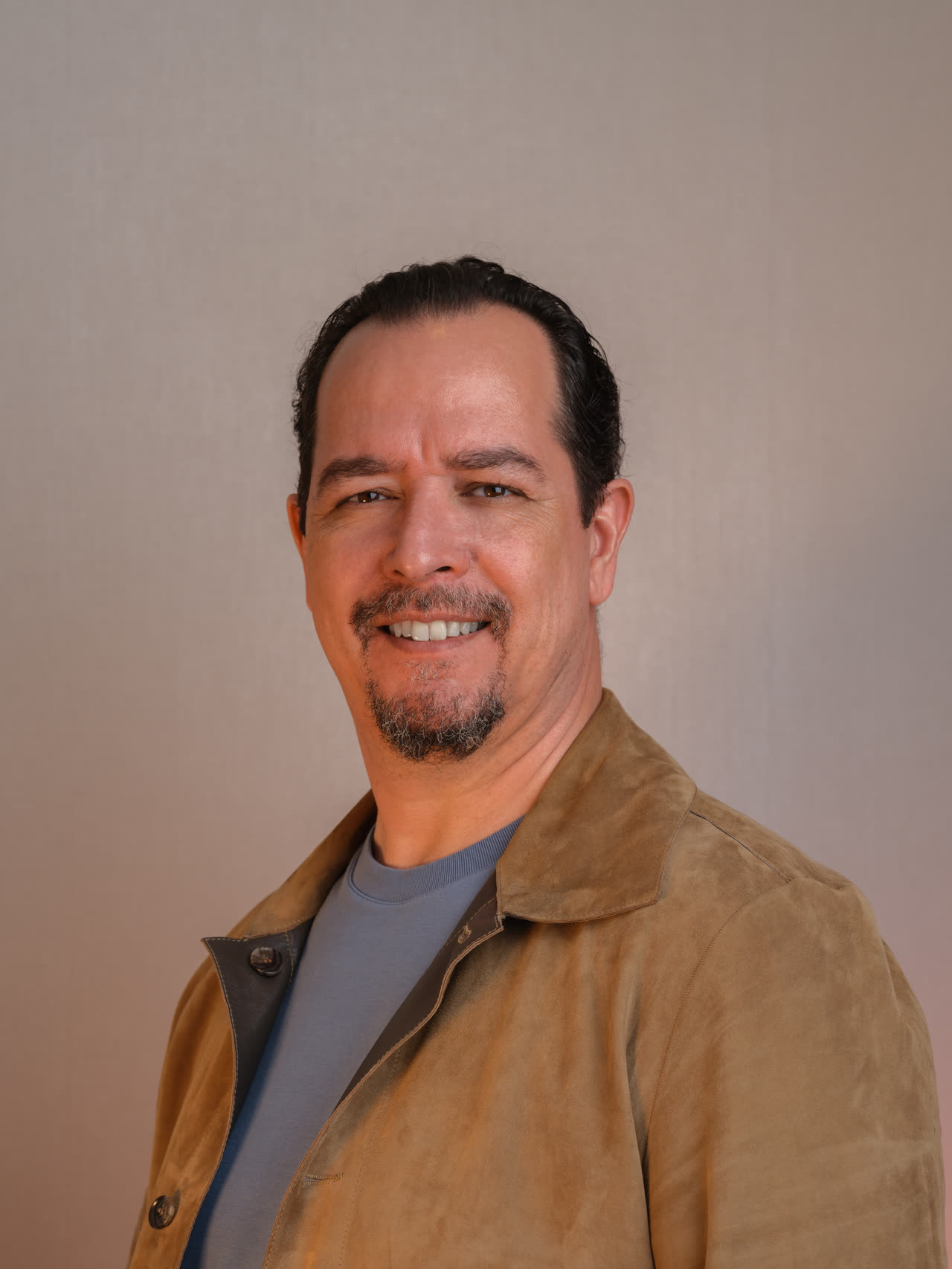 Portrait of Dr Atakan Elter, smiling to camera, hair swept back, greying beard; a suede jacket over a blue-grey t-shirt, warm neutral ground.